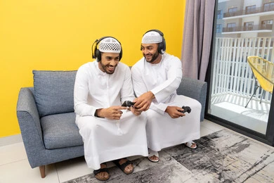 Using modern technology for fun and entertainment, playing video games at home, listening to music through headphones, two Emirati Gulf Arab men wearing kandoras and hats, putting on headphones and holding joysticks, spending quality time with friends
