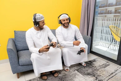نشاط شبابي ترفيهي، مفهوم المتعة والتسلية، اللعب بألعاب الفيديو الترفيهية مع الأصدقاء في المنزل، الاصغاء والاستماع عبر الهيدفون، استخدام الأجهزة التكنولوجية الحديثة، رجلان عربيان خليجيان اماراتيان يرتديان الكندورة والقبعة يضعان سماعات الرأس ويمسكان بيدهما عصا التحكم