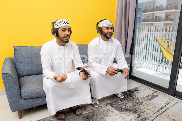 الاستمتاع والتسلية في العطلة مع الاصدقاء، استخدام التقنية الحديثة في المتعة والترفيه، اللعب بألعاب الفيديو الترفيهية في المنزل، الاصغاء والاستماع عبر الهيدفون، رجلان عربيان خليجيان اماراتيان يرتديان الكندورة والقبعة يضعان سماعات الرأس ويمسكان بيدهما عصا التحكم