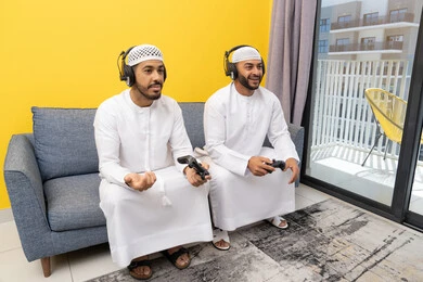 الاستمتاع والتسلية في العطلة مع الاصدقاء، استخدام التقنية الحديثة في المتعة والترفيه، اللعب بألعاب الفيديو الترفيهية في المنزل، الاصغاء والاستماع عبر الهيدفون، رجلان عربيان خليجيان اماراتيان يرتديان الكندورة والقبعة يضعان سماعات الرأس ويمسكان بيدهما عصا التحكم