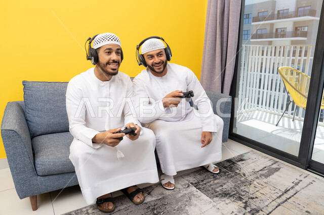 نشاط شبابي ترفيهي، مفهوم المتعة والتسلية، اللعب بألعاب الفيديو الترفيهية مع الأصدقاء في المنزل، الاصغاء والاستماع عبر الهيدفون، استخدام الأجهزة التكنولوجية الحديثة، رجلان عربيان خليجيان اماراتيان يرتديان الكندورة والقبعة يضعان سماعات الرأس ويمسكان بيدهما عصا التحكم