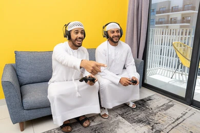 Using modern technology for fun and entertainment, playing video games at home, listening to music through headphones, two Emirati Gulf Arab men wearing kandoras and hats, putting on headphones and holding joysticks, spending quality time with friends