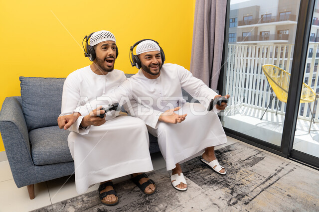 نشاط شبابي ترفيهي، مفهوم المتعة والتسلية، اللعب بألعاب الفيديو الترفيهية مع الأصدقاء في المنزل، الاصغاء والاستماع عبر الهيدفون، استخدام الأجهزة التكنولوجية الحديثة، رجلان عربيان خليجيان اماراتيان يرتديان الكندورة والقبعة يضعان سماعات الرأس ويمسكان بيدهما عصا التحكم