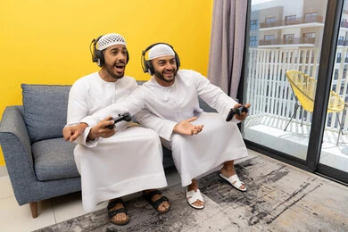 نشاط شبابي ترفيهي، مفهوم المتعة والتسلية، اللعب بألعاب الفيديو الترفيهية مع الأصدقاء في المنزل، الاصغاء والاستماع عبر الهيدفون، استخدام الأجهزة التكنولوجية الحديثة، رجلان عربيان خليجيان اماراتيان يرتديان الكندورة والقبعة يضعان سماعات الرأس ويمسكان بيدهما عصا التحكم