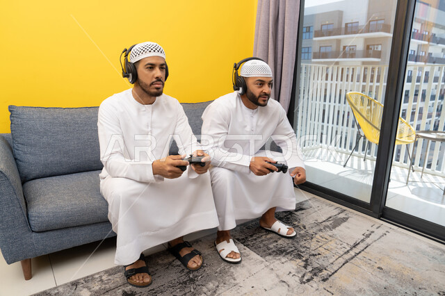 Using modern technology for fun and entertainment, playing video games at home, listening to music through headphones, two Emirati Gulf Arab men wearing kandoras and hats, putting on headphones and holding joysticks, spending quality time with friends