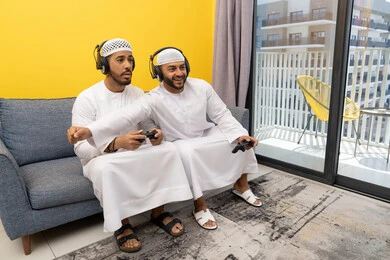الاستمتاع والتسلية في العطلة مع الاصدقاء، استخدام التقنية الحديثة في المتعة والترفيه، اللعب بألعاب الفيديو الترفيهية في المنزل، الاصغاء والاستماع عبر الهيدفون، رجلان عربيان خليجيان اماراتيان يرتديان الكندورة والقبعة يضعان سماعات الرأس ويمسكان بيدهما عصا التحكم