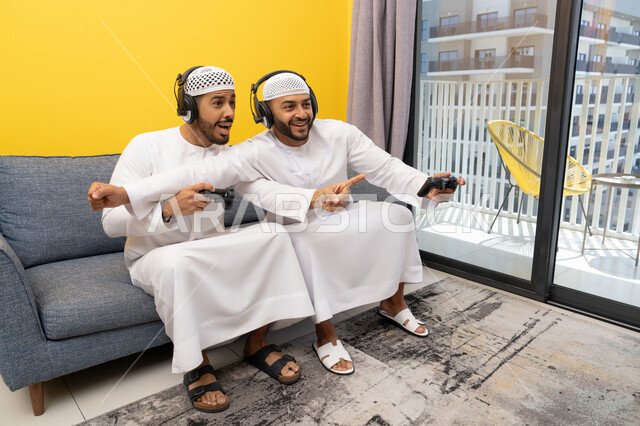 استخدام التقنية الحديثة في المتعة والترفيه، اللعب بألعاب الفيديو الترفيهية في المنزل، الاصغاء والاستماع عبر الهيدفون، رجلان عربيان خليجيان اماراتيان يرتديان الكندورة والقبعة يضعان سماعات الرأس ويمسكان بيدهما عصا التحكم، قضاء وقت ممتع مع الاصدقاء