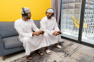 اللعب بألعاب الفيديو الترفيهية في المنزل، الاصغاء والاستماع عبر الهيدفون، رجلان عربيان خليجيان اماراتيان يرتديان الكندورة والقبعة يضعان سماعات الرأس ويمسكان بيدهما عصا التحكم، قضاء وقت ممتع مع الاصدقاء، المشاركة والمنافسة بالألعاب، نشاط شبابي ترفيهي