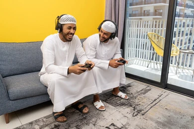 Playing recreational video games at home, listening and hearing through headphones, two Emirati Gulf Arab men wearing kandora and hat, putting on headphones and holding joysticks, spending a fun time with friends, participating and competing in games, a recreational youth activity