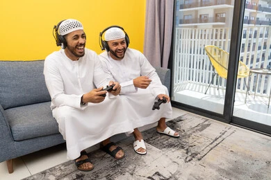 قضاء وقت ممتع مع الاصدقاء، المشاركة والمنافسة بالألعاب، نشاط شبابي ترفيهي، اللعب بألعاب الفيديو الترفيهية في المنزل، الاصغاء والاستماع عبر الهيدفون، رجلان عربيان خليجيان اماراتيان يرتديان الكندورة والقبعة يضعان سماعات الرأس ويمسكان بيدهما عصا التحكم
