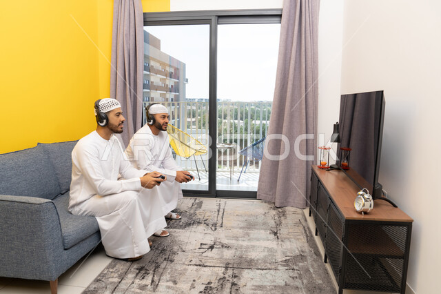 Recreational youth activity, the concept of fun and entertainment, playing recreational video games with friends at home, listening and hearing through headphones, using modern technological devices, two Emirati Gulf Arab men wearing kandoras and hats, putting on headphones and holding joysticks