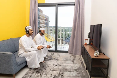 نشاط شبابي ترفيهي، مفهوم المتعة والتسلية، اللعب بألعاب الفيديو الترفيهية مع الأصدقاء في المنزل، الاصغاء والاستماع عبر الهيدفون، استخدام الأجهزة التكنولوجية الحديثة، رجلان عربيان خليجيان اماراتيان يرتديان الكندورة والقبعة يضعان سماعات الرأس ويمسكان بيدهما عصا التحكم