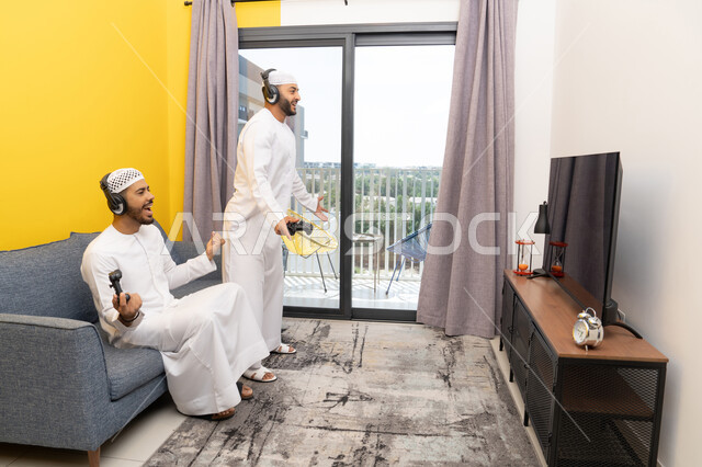 قضاء وقت ممتع مع الاصدقاء، المشاركة والمنافسة بالألعاب، نشاط شبابي ترفيهي، اللعب بألعاب الفيديو الترفيهية في المنزل، الاصغاء والاستماع عبر الهيدفون، رجلان عربيان خليجيان اماراتيان يرتديان الكندورة والقبعة يضعان سماعات الرأس ويمسكان بيدهما عصا التحكم