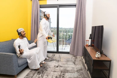 قضاء وقت ممتع مع الاصدقاء، المشاركة والمنافسة بالألعاب، نشاط شبابي ترفيهي، اللعب بألعاب الفيديو الترفيهية في المنزل، الاصغاء والاستماع عبر الهيدفون، رجلان عربيان خليجيان اماراتيان يرتديان الكندورة والقبعة يضعان سماعات الرأس ويمسكان بيدهما عصا التحكم