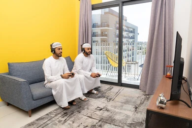 نشاط شبابي ترفيهي، مفهوم المتعة والتسلية، اللعب بألعاب الفيديو الترفيهية مع الأصدقاء في المنزل، الاصغاء والاستماع عبر الهيدفون، استخدام الأجهزة التكنولوجية الحديثة، رجلان عربيان خليجيان اماراتيان يرتديان الكندورة والقبعة يضعان سماعات الرأس ويمسكان بيدهما عصا التحكم