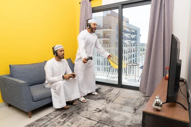 اللعب بألعاب الفيديو الترفيهية في المنزل، الاصغاء والاستماع عبر الهيدفون، رجلان عربيان خليجيان اماراتيان يرتديان الكندورة والقبعة يضعان سماعات الرأس ويمسكان بيدهما عصا التحكم، قضاء وقت ممتع مع الاصدقاء، المشاركة والمنافسة بالألعاب، نشاط شبابي ترفيهي