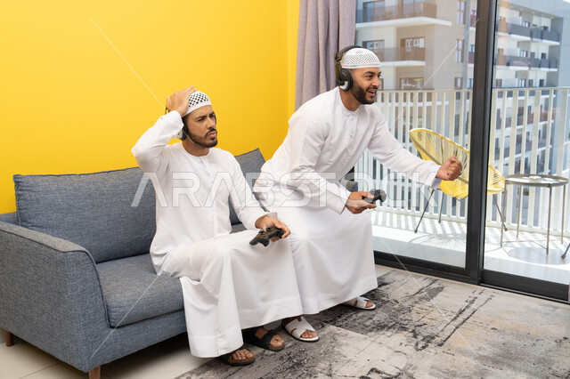قضاء وقت ممتع مع الاصدقاء، المشاركة والمنافسة بالألعاب، نشاط شبابي ترفيهي، اللعب بألعاب الفيديو الترفيهية في المنزل، الاصغاء والاستماع عبر الهيدفون، رجلان عربيان خليجيان اماراتيان يرتديان الكندورة والقبعة يضعان سماعات الرأس ويمسكان بيدهما عصا التحكم
