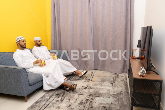 ايماءات الاندماج والسرور، مفهوم التسليه والترفيه الجماعي, قضاء وقت ممتع بتناول الفوشار اثناء مشاهده التلفاز، صديقان عربيان خليجيان اماراتيان يرتديان الكندورة والطاقية البيضاء يجلسان على الأريكه يتناولان البوب كورن، الاستمتاع بالأنشطة الترفيهيه في عطلة نهاية الاسبوع
