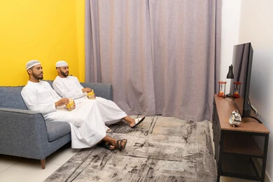 ايماءات الاندماج والسرور، مفهوم التسليه والترفيه الجماعي, قضاء وقت ممتع بتناول الفوشار اثناء مشاهده التلفاز، صديقان عربيان خليجيان اماراتيان يرتديان الكندورة والطاقية البيضاء يجلسان على الأريكه يتناولان البوب كورن، الاستمتاع بالأنشطة الترفيهيه في عطلة نهاية الاسبوع