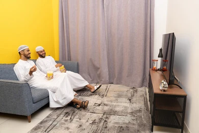 Youth entertainment and amusement, having a good time eating popcorn while watching TV, two Emirati Gulf Arab friends wearing kanduras and white taqiyas sitting on the couch eating popcorn, enjoying recreational activities on the weekend, gestures of integration and pleasure