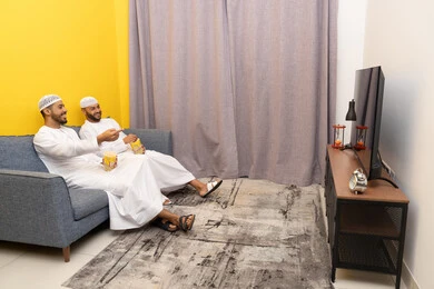 مفهوم التسليه والترفيه الجماعي, قضاء وقت ممتع بتناول الفوشار اثناء مشاهده التلفاز، صديقان عربيان خليجيان اماراتيان يرتديان الكندورة والطاقية البيضاء يجلسان على الأريكه يتناولان البوب كورن، الاستمتاع بالأنشطة الترفيهيه في عطلة نهاية الاسبوع، ايماءات الاندماج والسرور