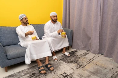 مفهوم التسليه والترفيه الجماعي, قضاء وقت ممتع بتناول الفوشار اثناء مشاهده التلفاز، صديقان عربيان خليجيان اماراتيان يرتديان الكندورة والطاقية البيضاء يجلسان على الأريكه يتناولان البوب كورن، الاستمتاع بالأنشطة الترفيهيه في عطلة نهاية الاسبوع، ايماءات الاندماج والسرور