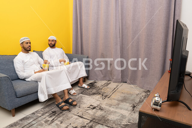 التسليه والترفيه الشبابي, قضاء وقت ممتع بتناول الفوشار اثناء مشاهده التلفاز، صديقان عربيان خليجيان اماراتيان يرتديان الكندورة والطاقية البيضاء يجلسان على الأريكه يتناولان البوب كورن، الاستمتاع بالأنشطة الترفيهيه في عطلة نهاية الاسبوع، ايماءات الاندماج والسرور
