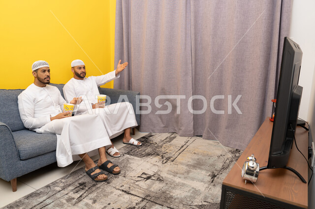 ايماءات الاندماج والسرور، مفهوم التسليه والترفيه الجماعي, قضاء وقت ممتع بتناول الفوشار اثناء مشاهده التلفاز، صديقان عربيان خليجيان اماراتيان يرتديان الكندورة والطاقية البيضاء يجلسان على الأريكه يتناولان البوب كورن، الاستمتاع بالأنشطة الترفيهيه في عطلة نهاية الاسبوع