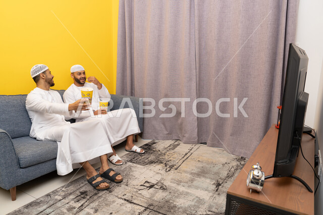 مفهوم التسليه والترفيه الجماعي, قضاء وقت ممتع بتناول الفوشار اثناء مشاهده التلفاز، صديقان عربيان خليجيان اماراتيان يرتديان الكندورة والطاقية البيضاء يجلسان على الأريكه يتناولان البوب كورن، الاستمتاع بالأنشطة الترفيهيه في عطلة نهاية الاسبوع، ايماءات الاندماج والسرور