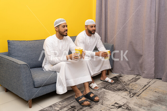 ايماءات الاندماج والسرور، مفهوم التسليه والترفيه الجماعي, قضاء وقت ممتع بتناول الفوشار اثناء مشاهده التلفاز، صديقان عربيان خليجيان اماراتيان يرتديان الكندورة والطاقية البيضاء يجلسان على الأريكه يتناولان البوب كورن، الاستمتاع بالأنشطة الترفيهيه في عطلة نهاية الاسبوع