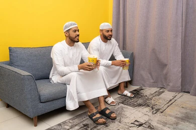 ايماءات الاندماج والسرور، مفهوم التسليه والترفيه الجماعي, قضاء وقت ممتع بتناول الفوشار اثناء مشاهده التلفاز، صديقان عربيان خليجيان اماراتيان يرتديان الكندورة والطاقية البيضاء يجلسان على الأريكه يتناولان البوب كورن، الاستمتاع بالأنشطة الترفيهيه في عطلة نهاية الاسبوع