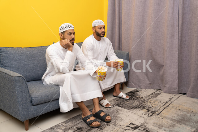 الترفيه والتسلية الشبابي, قضاء وقت ممتع بتناول الفوشار اثناء مشاهده التلفاز، صديقان عربيان خليجيان اماراتيان يرتديان الكندورة والطاقية البيضاء يجلسان على الأريكه يتناولان البوب كورن، الاستمتاع بالأنشطة الترفيهيه في عطلة نهاية الاسبوع، ايماءات الاندماج والسرور