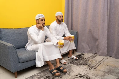 الترفيه والتسلية الشبابي, قضاء وقت ممتع بتناول الفوشار اثناء مشاهده التلفاز، صديقان عربيان خليجيان اماراتيان يرتديان الكندورة والطاقية البيضاء يجلسان على الأريكه يتناولان البوب كورن، الاستمتاع بالأنشطة الترفيهيه في عطلة نهاية الاسبوع، ايماءات الاندماج والسرور