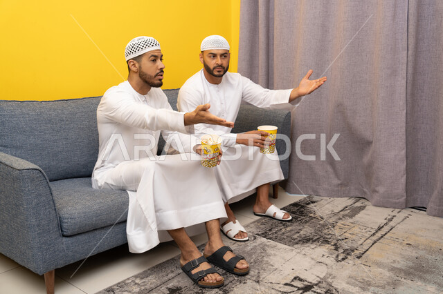 مفهوم التسليه والترفيه الجماعي, قضاء وقت ممتع بتناول الفوشار اثناء مشاهده التلفاز، صديقان عربيان خليجيان اماراتيان يرتديان الكندورة والطاقية البيضاء يجلسان على الأريكه يتناولان البوب كورن، الاستمتاع بالأنشطة الترفيهيه في عطلة نهاية الاسبوع، ايماءات الاندماج والسرور
