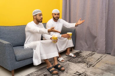 مفهوم التسليه والترفيه الجماعي, قضاء وقت ممتع بتناول الفوشار اثناء مشاهده التلفاز، صديقان عربيان خليجيان اماراتيان يرتديان الكندورة والطاقية البيضاء يجلسان على الأريكه يتناولان البوب كورن، الاستمتاع بالأنشطة الترفيهيه في عطلة نهاية الاسبوع، ايماءات الاندماج والسرور