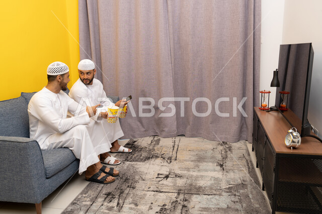 التسليه والترفيه الشبابي, قضاء وقت ممتع بتناول الفوشار اثناء مشاهده التلفاز، صديقان عربيان خليجيان اماراتيان يرتديان الكندورة والطاقية البيضاء يجلسان على الأريكه يتناولان البوب كورن، الاستمتاع بالأنشطة الترفيهيه في عطلة نهاية الاسبوع، ايماءات الاندماج والسرور