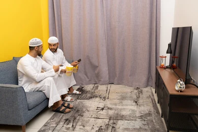 التسليه والترفيه الشبابي, قضاء وقت ممتع بتناول الفوشار اثناء مشاهده التلفاز، صديقان عربيان خليجيان اماراتيان يرتديان الكندورة والطاقية البيضاء يجلسان على الأريكه يتناولان البوب كورن، الاستمتاع بالأنشطة الترفيهيه في عطلة نهاية الاسبوع، ايماءات الاندماج والسرور