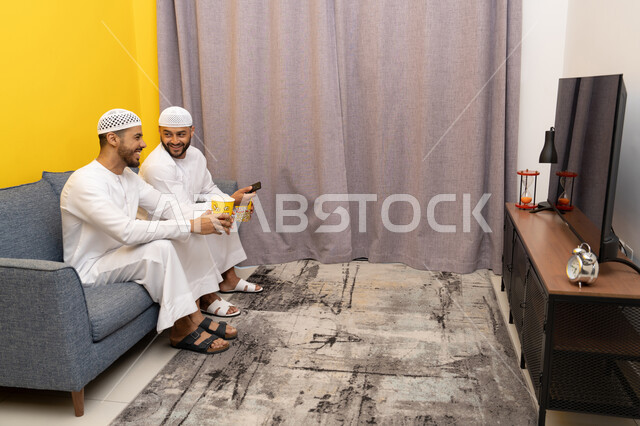 ايماءات الاندماج والسرور، مفهوم التسليه والترفيه الجماعي, قضاء وقت ممتع بتناول الفوشار اثناء مشاهده التلفاز، صديقان عربيان خليجيان اماراتيان يرتديان الكندورة والطاقية البيضاء يجلسان على الأريكه يتناولان البوب كورن، الاستمتاع بالأنشطة الترفيهيه في عطلة نهاية الاسبوع