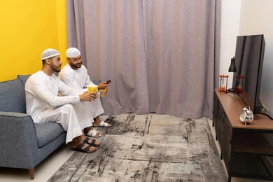 مفهوم التسليه والترفيه الجماعي, قضاء وقت ممتع بتناول الفوشار اثناء مشاهده التلفاز، صديقان عربيان خليجيان اماراتيان يرتديان الكندورة والطاقية البيضاء يجلسان على الأريكه يتناولان البوب كورن، الاستمتاع بالأنشطة الترفيهيه في عطلة نهاية الاسبوع، ايماءات الاندماج والسرور