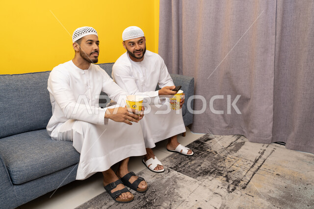 التسليه والترفيه الشبابي, قضاء وقت ممتع بتناول الفوشار اثناء مشاهده التلفاز، صديقان عربيان خليجيان اماراتيان يرتديان الكندورة والطاقية البيضاء يجلسان على الأريكه يتناولان البوب كورن، الاستمتاع بالأنشطة الترفيهيه في عطلة نهاية الاسبوع، ايماءات الاندماج والسرور