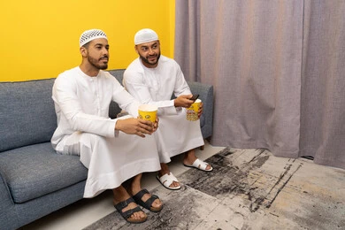 التسليه والترفيه الشبابي, قضاء وقت ممتع بتناول الفوشار اثناء مشاهده التلفاز، صديقان عربيان خليجيان اماراتيان يرتديان الكندورة والطاقية البيضاء يجلسان على الأريكه يتناولان البوب كورن، الاستمتاع بالأنشطة الترفيهيه في عطلة نهاية الاسبوع، ايماءات الاندماج والسرور