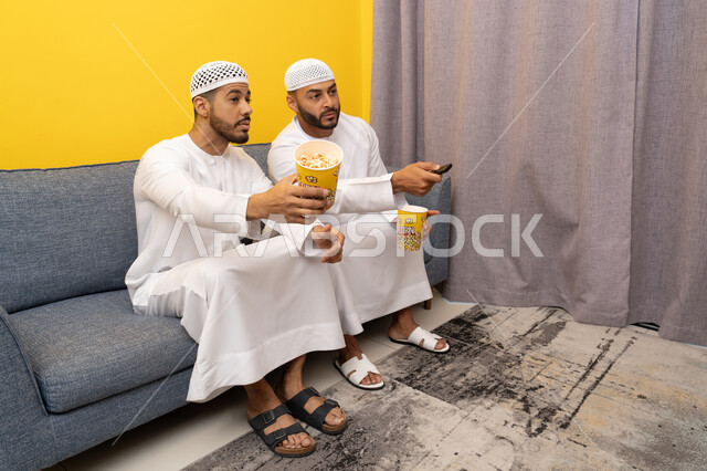 ايماءات الاندماج والسرور، مفهوم التسليه والترفيه الجماعي, قضاء وقت ممتع بتناول الفوشار اثناء مشاهده التلفاز، صديقان عربيان خليجيان اماراتيان يرتديان الكندورة والطاقية البيضاء يجلسان على الأريكه يتناولان البوب كورن، الاستمتاع بالأنشطة الترفيهيه في عطلة نهاية الاسبوع