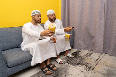 ايماءات الاندماج والسرور، مفهوم التسليه والترفيه الجماعي, قضاء وقت ممتع بتناول الفوشار اثناء مشاهده التلفاز، صديقان عربيان خليجيان اماراتيان يرتديان الكندورة والطاقية البيضاء يجلسان على الأريكه يتناولان البوب كورن، الاستمتاع بالأنشطة الترفيهيه في عطلة نهاية الاسبوع