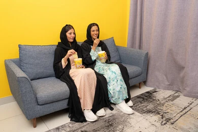 Enjoying popcorn while watching TV, spending quality time with friends, two Emirati Gulf Arab women wearing abaya and hijab sitting on the couch watching an entertaining movie with gestures of integration and pleasure, enjoying free time, a group home entertainment activity
