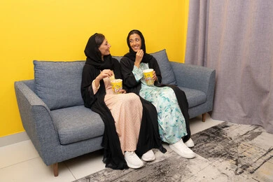 الاستمتاع باوقات الفراغ, نشاط منزلي ترفيهي جماعي, المتعة في تناول الفشار اثناء مشاهدة التلفاز، قضاء اوقات ممتعة بين الاصدقاء, إمرأتان عربيتان خليجيتان اماراتيان ترتديان العباءه والحجاب تجلسان على الكنبة تشاهدان فيلم مسلي بايماءات الاندماج و السرور