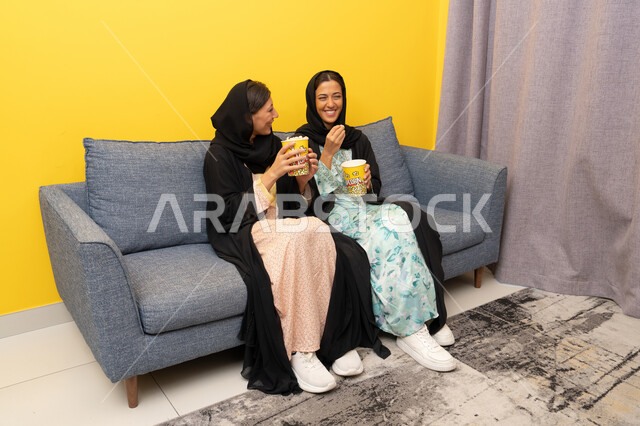 Enjoying popcorn while watching TV, spending quality time with friends, two Emirati Gulf Arab women wearing abaya and hijab sitting on the couch watching an entertaining movie with gestures of integration and pleasure, enjoying free time, a group home entertainment activity