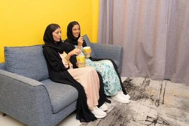 قضاء اوقات ممتعة بين الاصدقاء, إمرأتان عربيتان خليجيتان اماراتيان ترتديان العباءه والحجاب تجلسان على الكنبة تشاهدان فيلم مسلي بايماءات الاندماج و السرور، الاستمتاع باوقات الفراغ، نشاط منزلي ترفيهي جماعي، المتعة في تناول الفشار اثناء مشاهدة التلفاز