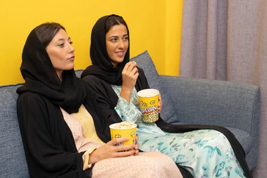 Two Emirati Gulf Arab women wearing abaya and hijab sitting on the sofa watching an entertaining movie with gestures of integration and pleasure, enjoying free time, a group recreational home activity, having fun eating popcorn while watching TV, spending quality time with friends