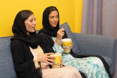 نشاط منزلي ترفيهي جماعي, المتعة في تناول الفشار اثناء مشاهدة التلفاز، قضاء اوقات ممتعة بين الاصدقاء, إمرأتان عربيتان خليجيتان اماراتيان ترتديان العباءه والحجاب تجلسان على الكنبة تشاهدان فيلم مسلي بايماءات الاندماج و السرور، الاستمتاع باوقات الفراغ