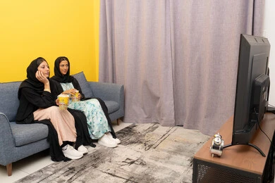 قضاء اوقات ممتعة بين الاصدقاء, إمرأتان عربيتان خليجيتان اماراتيان ترتديان العباءه والحجاب تجلسان على الكنبة تشاهدان فيلم مسلي بايماءات الاندماج و السرور، الاستمتاع باوقات الفراغ، نشاط منزلي ترفيهي جماعي، المتعة في تناول الفشار اثناء مشاهدة التلفاز